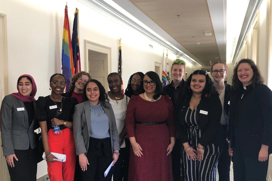 a group of students gather with a state representative in the. halls of a public building. The American flag and pride flag are directly behind them in the hallway.